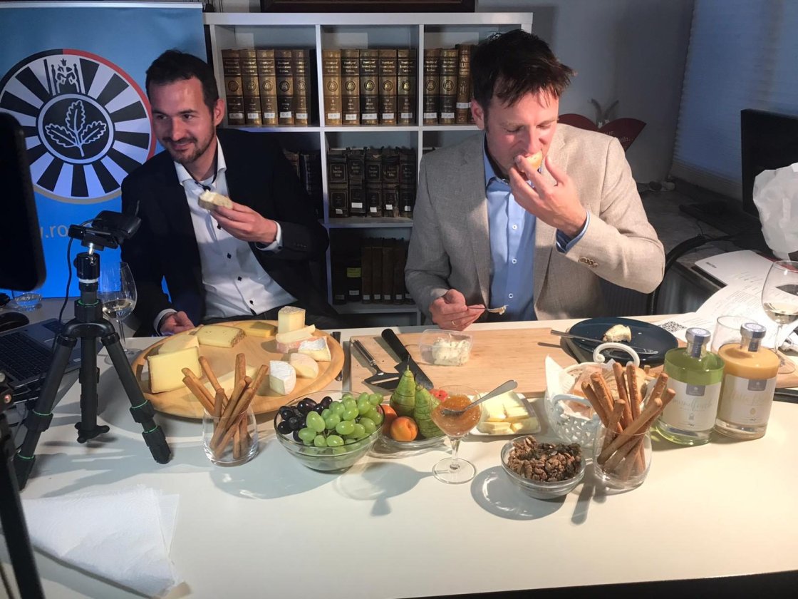 Zwei Gastgeber verkosten vor der Kamera Käse, auf dem Tisch eine Käseplatte mit Trauben und Grissini.