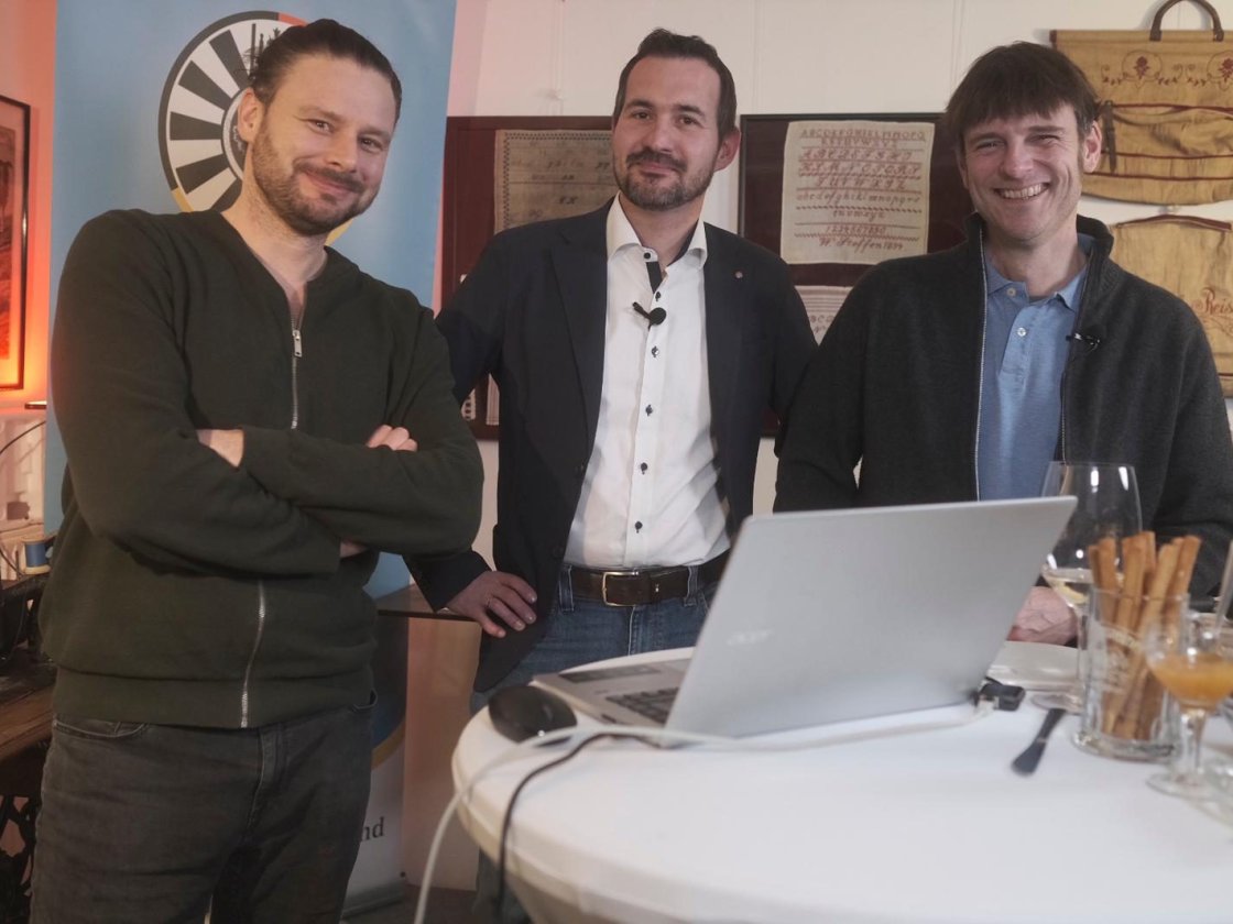 Drei Gastgeber stehen hinter dem Tisch mit Laptop und Käse, im Hintergrund das Round-Table-Banner.
