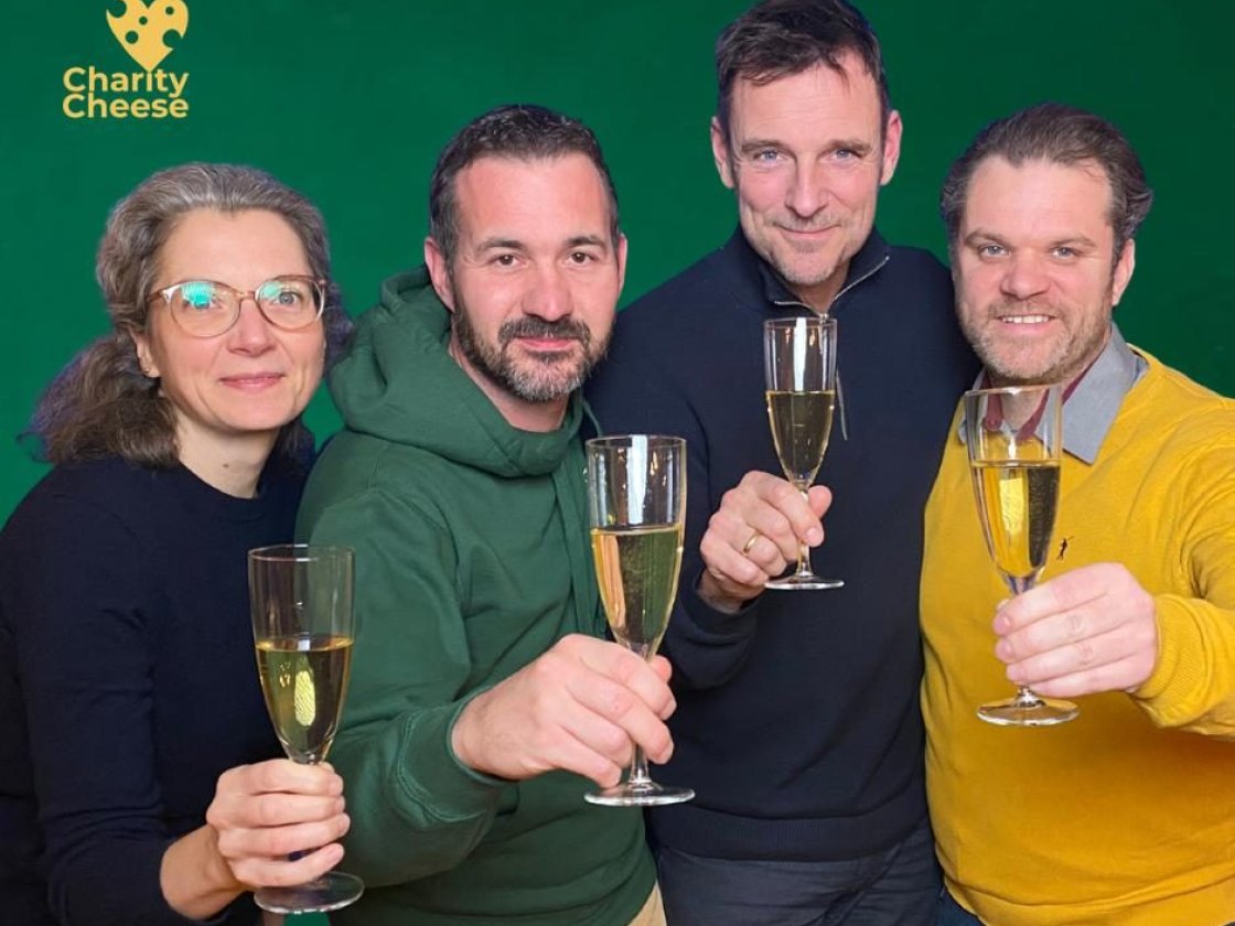 Vier Mitglieder des Charity Cheese e.V. stoßen vor grünem Hintergrund mit Sekt an, oben links das Logo.