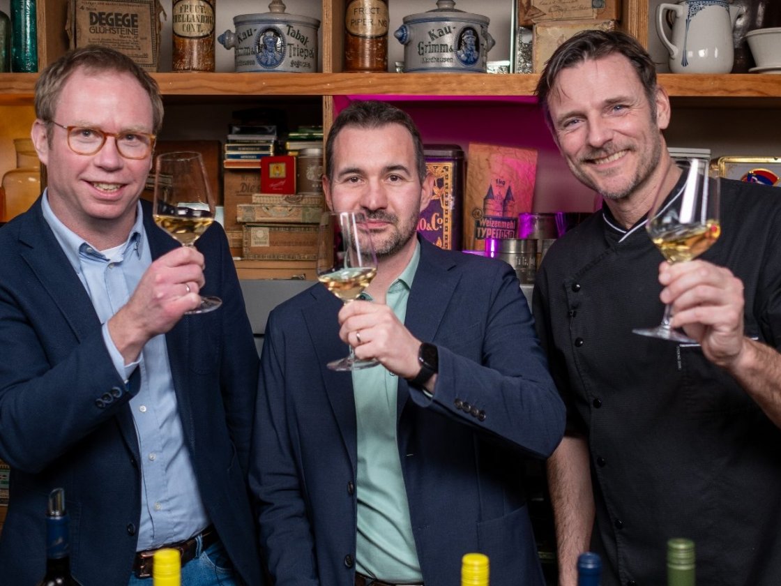 Drei Gastgeber stoßen im Kaufmannsladen mit Weißwein an.